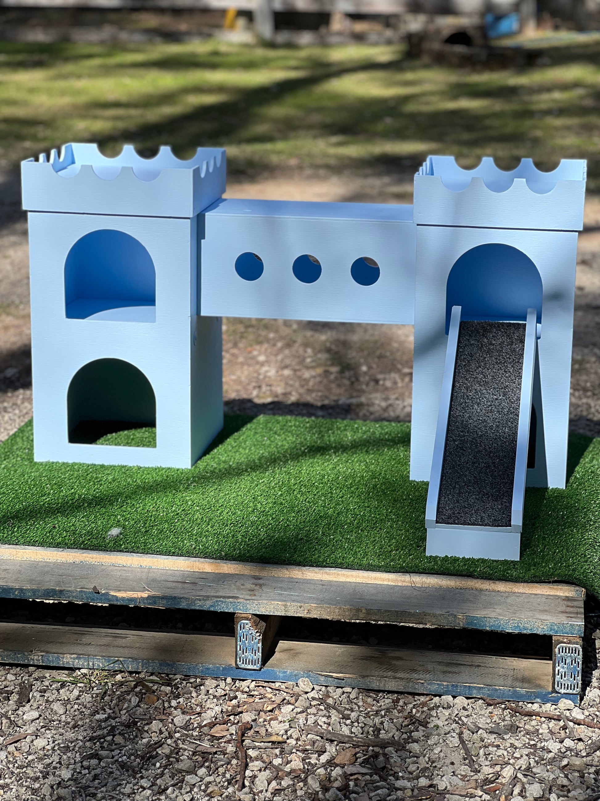 A small castle-themed outdoor play structure made of light blue wood, with arched openings, circular cutouts, a ramp, and a green artificial turf base, photographed outdoors on a wooden pallet over gravel.